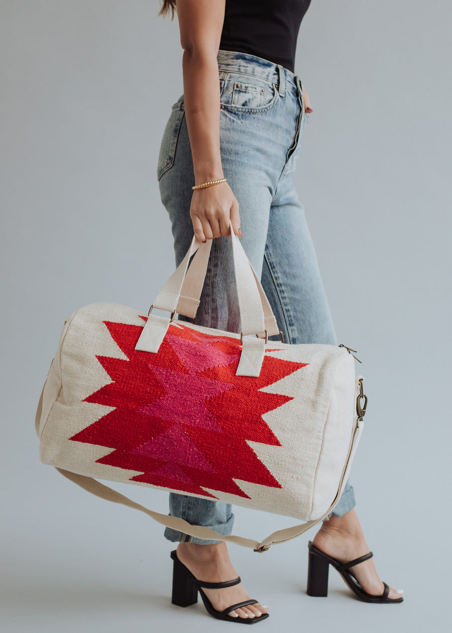 A person in jeans, a black top, and heeled sandals holds the panacheaccessories Stella Duffel—a cream-colored, western-inspired bag with red geometric patterns, cotton lining, handles, shoulder strap, gold hardware, and zipper closure. Plain gray background.