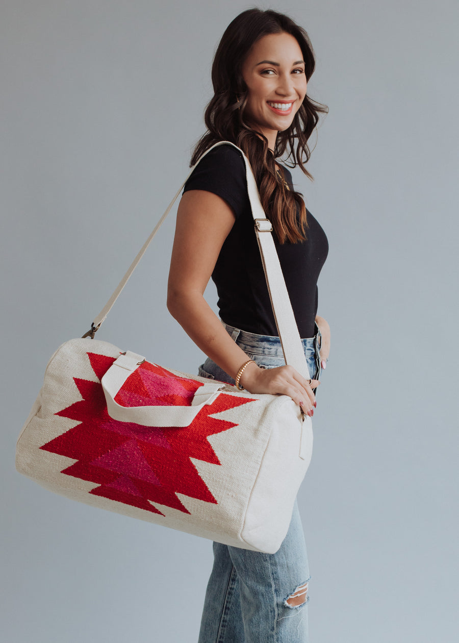 A smiling woman with long brown hair, in a black t-shirt and ripped blue jeans, stands sideways holding the Stella Duffel by panacheaccessories, featuring pink and red geometric patterns and a cotton lining, against a plain light background.