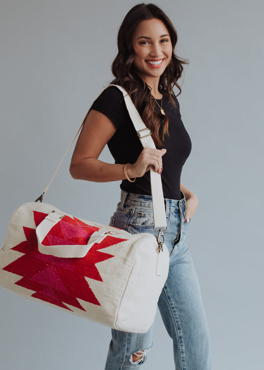 A woman with long brown hair, in a black t-shirt and ripped jeans, smiles while holding the panacheaccessories Stella Duffel—a western-inspired bag with a bold red geometric pattern and zipper closure—over her shoulder against a light gray background.