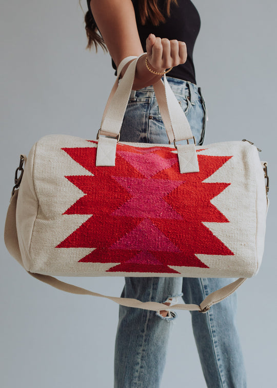 A woman in a black top and ripped jeans holds the panacheaccessories Stella Duffel, featuring bold red and pink geometric patterns, cream handles, a shoulder strap, and a secure zipper closure.