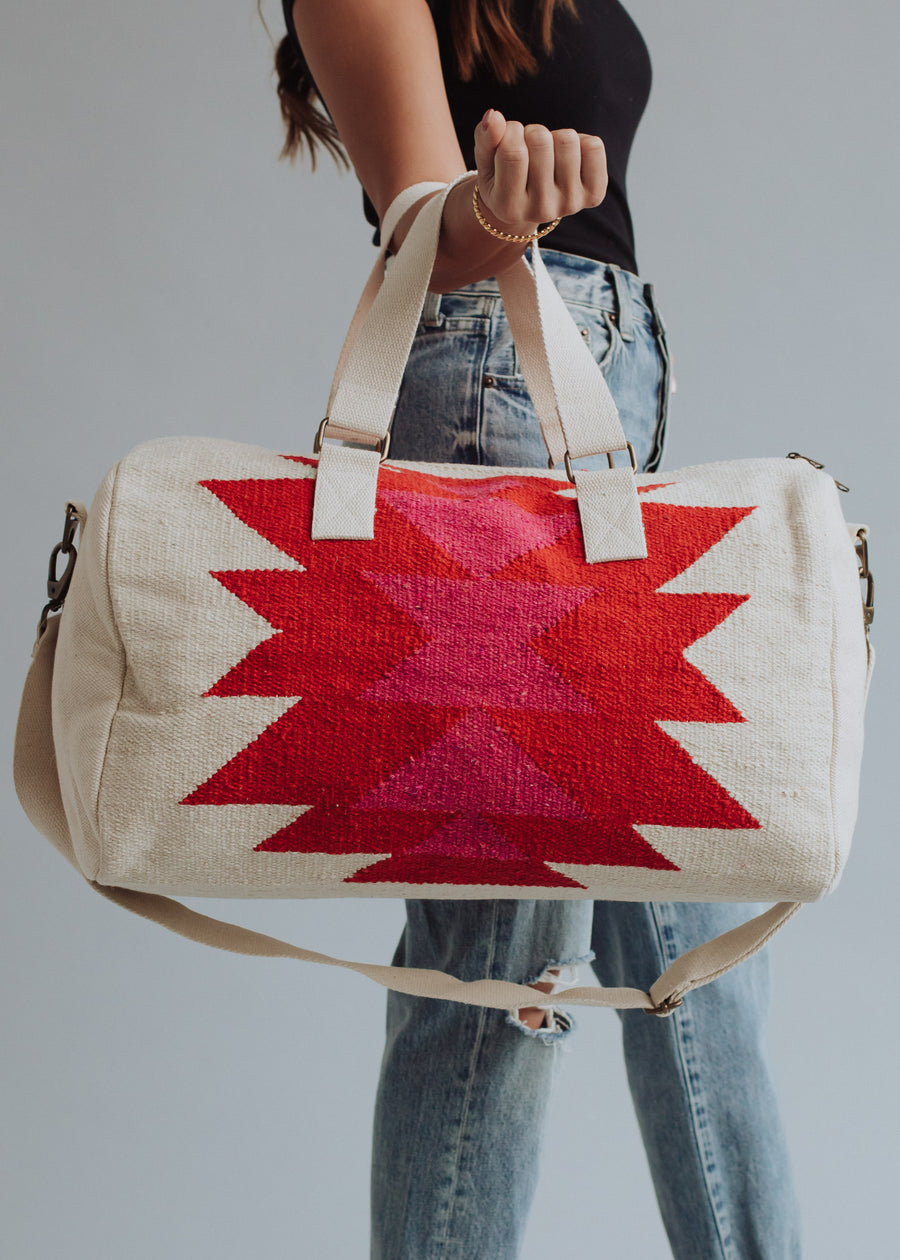 A woman in a black top and ripped jeans holds the panacheaccessories Stella Duffel, featuring bold red and pink geometric patterns, cream handles, a shoulder strap, and a secure zipper closure.
