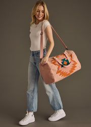 A smiling woman wearing a white t-shirt, blue jeans, and white sneakers carries the panacheaccessories Adalene Duffel, featuring a colorful geometric pattern and zipper closure, over her shoulder against a plain dark background.