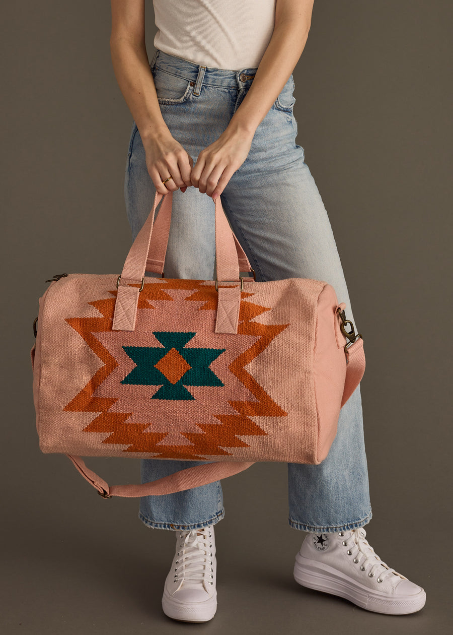 Person in light blue jeans, white shirt, and white high-top sneakers holds the panacheaccessories Adalene Duffel—a pink western bag with orange and green geometric pattern and zipper closure—against a plain background.