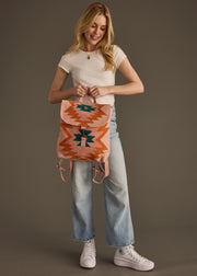 A smiling woman with long blonde hair holds the Adalene Backpack by panacheaccessories, featuring adjustable straps, while wearing a white t-shirt, light blue jeans, and white sneakers against a plain brown background.