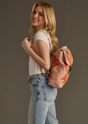 A woman with long blonde hair, wearing a white t-shirt and blue jeans, smiles while carrying the Adalene Backpack by panacheaccessories over her shoulder against a plain background.