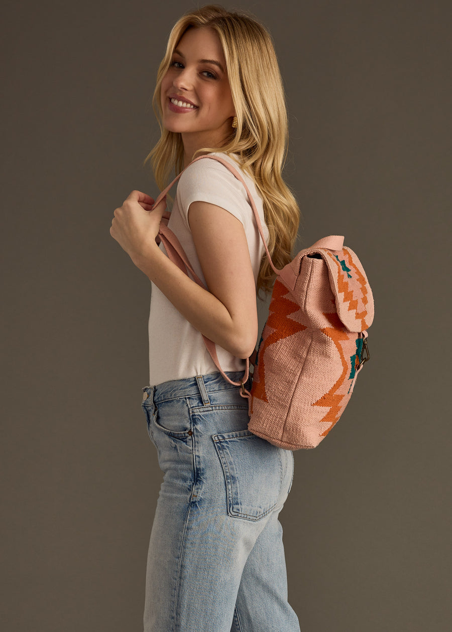 A woman with long blonde hair, wearing a white t-shirt and blue jeans, smiles while carrying the Adalene Backpack by panacheaccessories over her shoulder against a plain background.