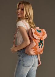 A smiling woman with long blonde hair stands against a dark background, wearing a white t-shirt, light blue jeans, and the Adalene Backpack by panacheaccessories—pink with orange and turquoise geometric patterns and adjustable straps.