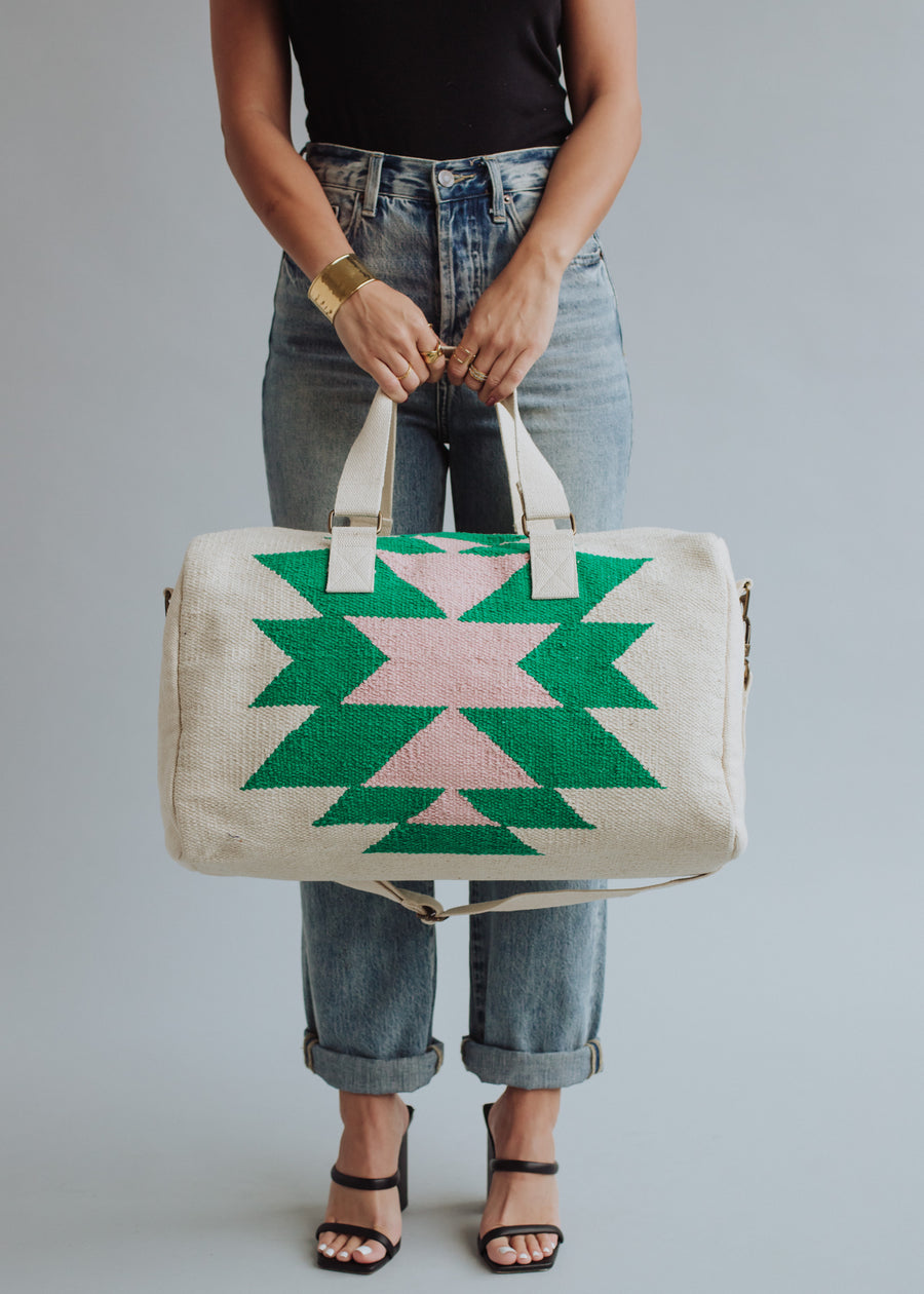 A person in a black top, jeans, and black heels holds the "One For Me Duffel" by panacheaccessories—a large cream cotton bag with pink and green geometric patterns and a zipper closure—against a plain gray background.