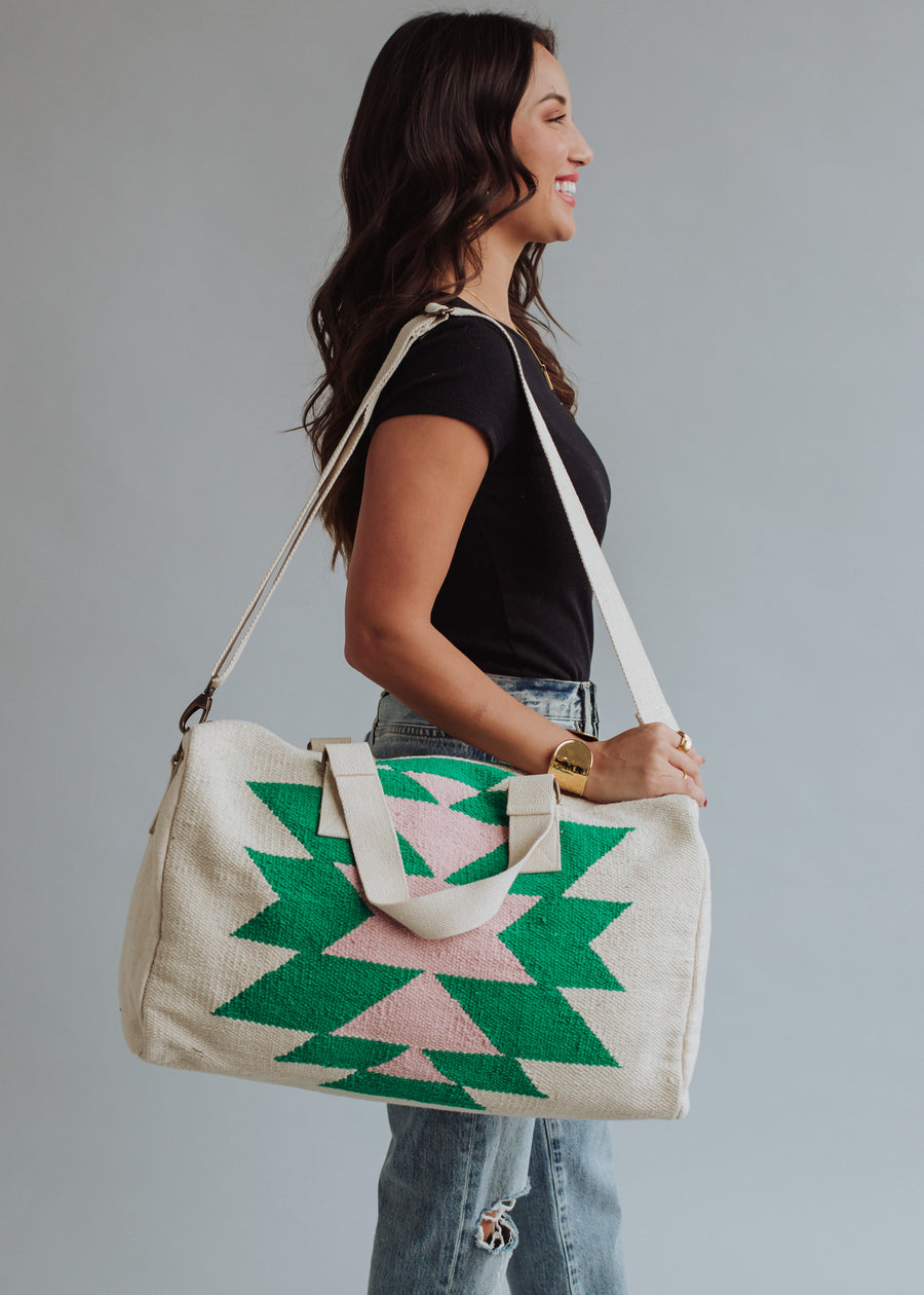 A woman in a black t-shirt and jeans smiles in profile, carrying the panacheaccessories One For Me Duffel—a large cream cotton bag with green and pink geometric patterns and zipper closure—over her shoulder against a plain background.