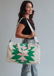 A woman in a black tee and ripped jeans smiles while holding the One For Me Duffel by panacheaccessories, a large cotton bag with zipper closure in green and pink geometric patterns, standing against a plain light gray background.