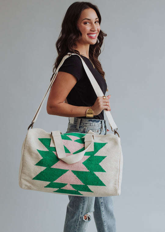 A woman in a black tee and ripped jeans smiles while holding the One For Me Duffel by panacheaccessories, a large cotton bag with zipper closure in green and pink geometric patterns, standing against a plain light gray background.