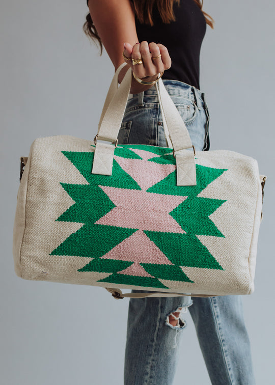 A person in ripped jeans and a black top holds the "One For Me Duffel" by panacheaccessories, a cream cotton bag with green and pink geometric patterns, beige handles, and a zipper closure against a plain background.