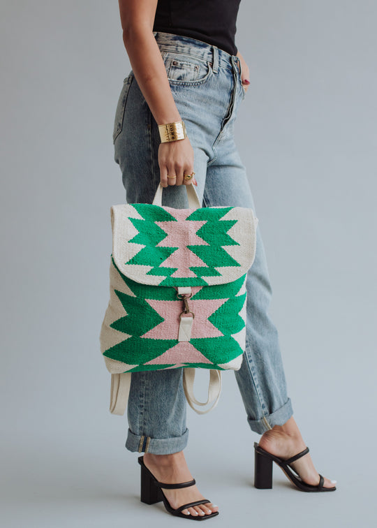 A person in cuffed jeans, black heels, and a black top holds the panacheaccessories One For Me Backpack, featuring adjustable straps and a bold green and pink geometric pattern, against a plain background.