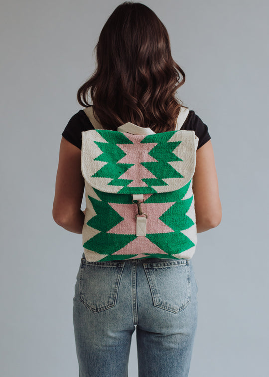 A woman with wavy brown hair, dressed in a black t-shirt and light blue jeans, faces away while carrying the panacheaccessories One For Me Backpack featuring a green, pink, and cream geometric western-inspired design. The background is plain light gray.