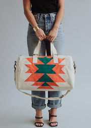 A person in jeans, black top, and sandals holds the Selkie Duffel by panacheaccessories. The beige, western-inspired bag has orange and green geometric patterns, short handles, a shoulder strap, and a secure zipper closure. The person's head is not visible.