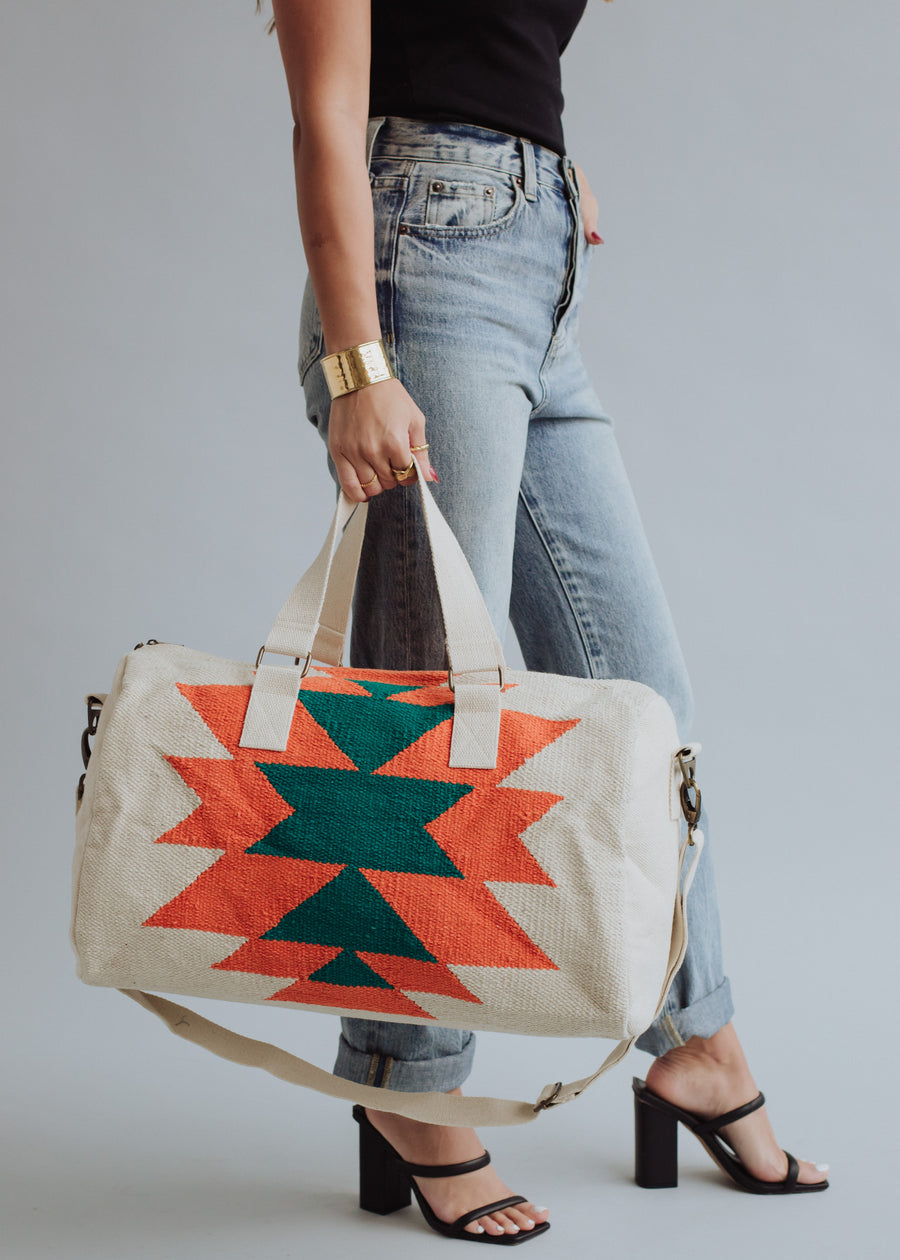 A woman in a black sleeveless top, light blue jeans, and black heeled sandals holds the panacheaccessories Selkie Duffel, a cream-colored bag with bold orange and green geometric patterns. She accessorizes with a gold cuff bracelet.