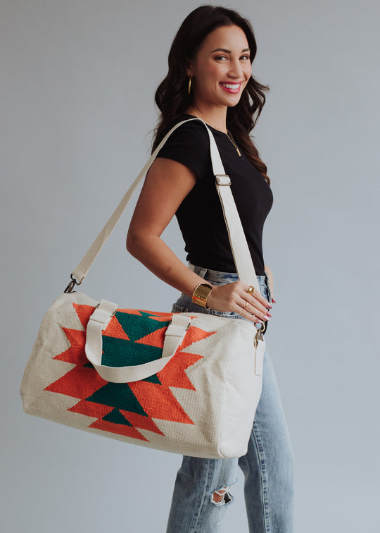 A woman in a black t-shirt and ripped jeans smiles while holding the panacheaccessories Selkie Duffel, a cream western-inspired bag with bold red and green geometric patterns, cotton lining, zipper closure, two handles, and a shoulder strap.