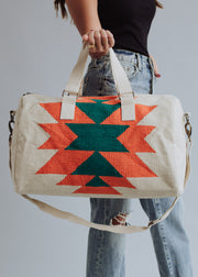 A person in a black top and ripped blue jeans holds the panacheaccessories Selkie Duffel, featuring a cream western-inspired design with bold orange and green geometric patterns, beige handles, a shoulder strap, zipper closure, and soft cotton lining.