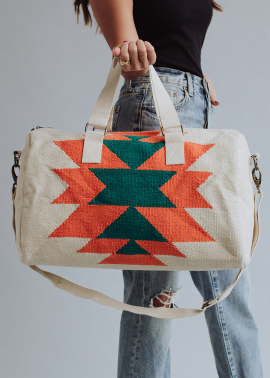 A person in a black top and ripped blue jeans holds the panacheaccessories Selkie Duffel, featuring a cream western-inspired design with bold orange and green geometric patterns, beige handles, a shoulder strap, zipper closure, and soft cotton lining.