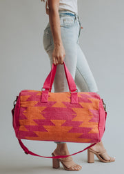 A person in light-wash jeans and heeled sandals holds the panacheaccessories Daybreak Duffel, a large pink and orange bag with handles, a shoulder strap, secure zipper closure, and a bold geometric design.