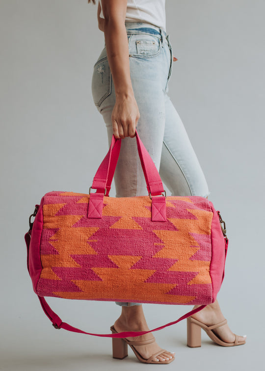 A person in light-wash jeans and heeled sandals holds the panacheaccessories Daybreak Duffel, a large pink and orange bag with handles, a shoulder strap, secure zipper closure, and a bold geometric design.