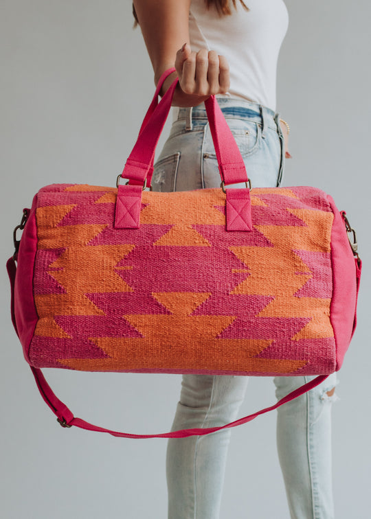 A woman in a white top and light jeans holds the Daybreak Duffel by panacheaccessories, featuring pink handles, an orange and pink geometric pattern, zipper closure, and a detachable shoulder strap.