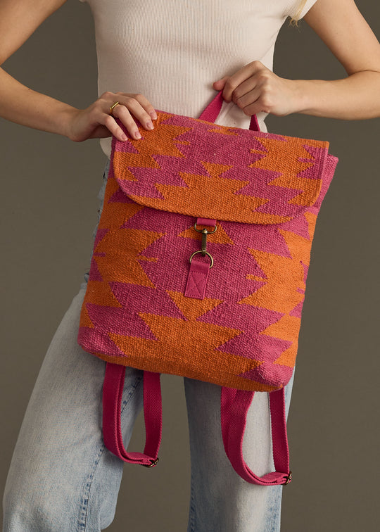 A person in a white sleeveless top and light jeans holds the panacheaccessories Daybreak Backpack, featuring pink and orange zigzag patterns, pink adjustable straps, and a clasp closure, against a neutral background.