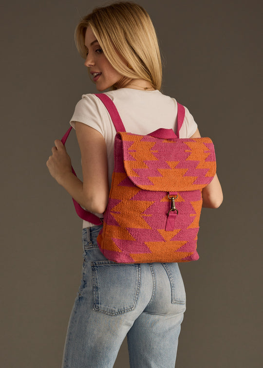 A woman in a white tee and blue jeans poses with the Daybreak Backpack by panacheaccessories, featuring a pink and orange western-inspired pattern and adjustable straps, smiling over her shoulder against a plain background.