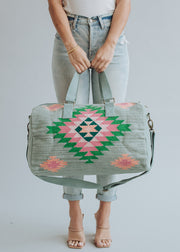 A woman in cuffed light-wash jeans and a white top carries the panacheaccessories Hadley Duffel, featuring green, pink, and black geometric patterns and a secure zipper closure. She pairs it with nude heeled sandals and a gold bracelet.