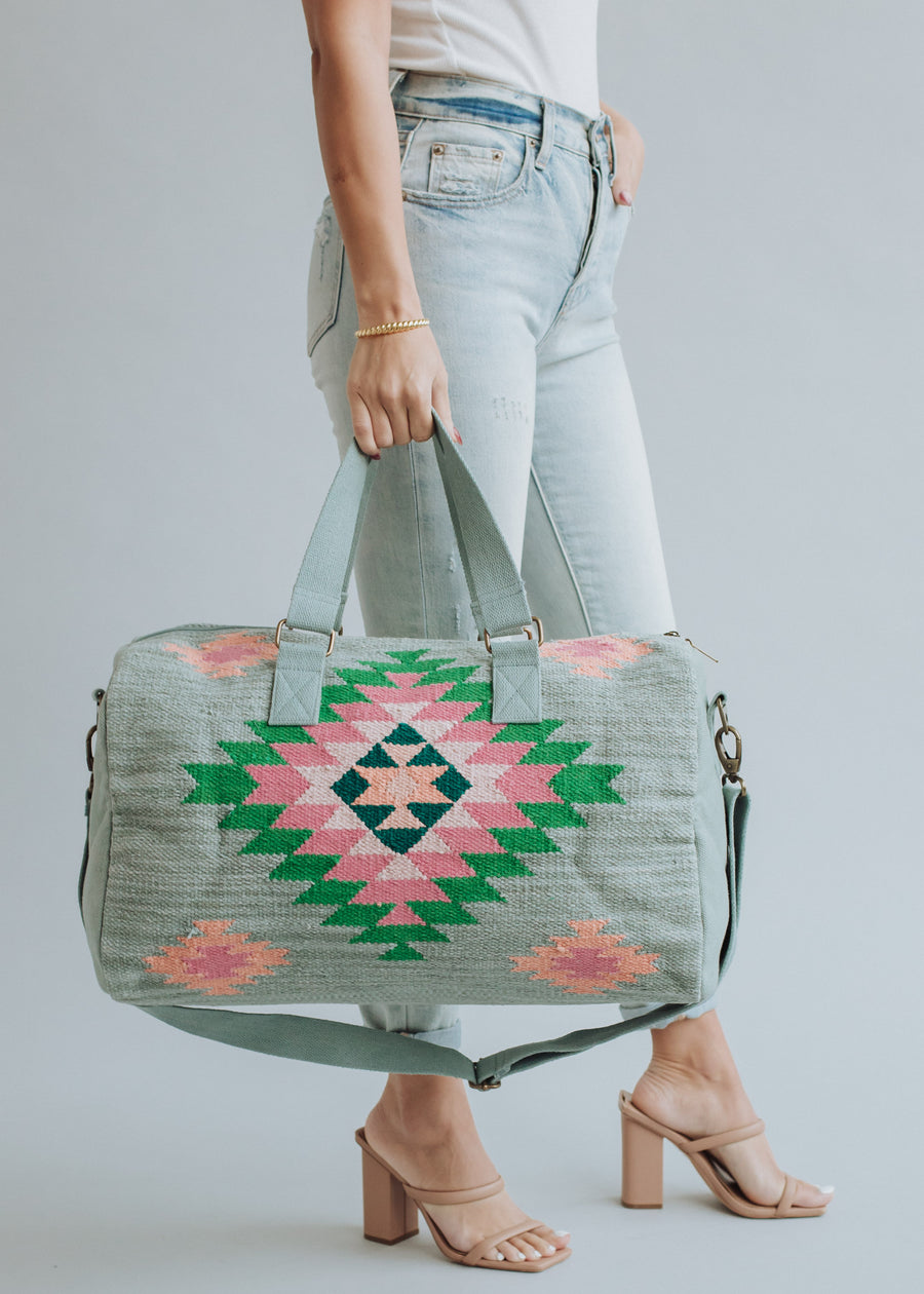 A person in light blue jeans and nude heels holds the panacheaccessories Hadley Duffel, which features a pink and green geometric pattern and secure zipper closure. The background is plain light gray.