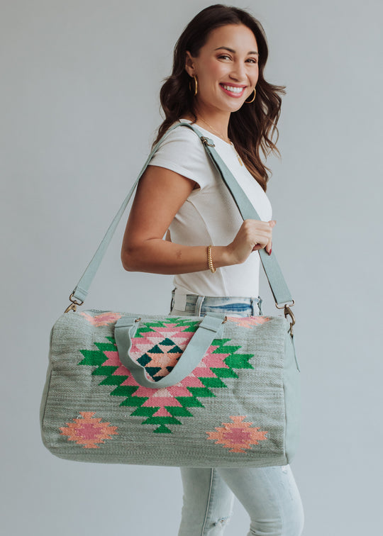 A smiling woman with long brown hair, in a white t-shirt and light jeans, carries the panacheaccessories Hadley Duffel—a large weekender bag with green, pink, and peach geometric designs and a secure zipper—over her shoulder.