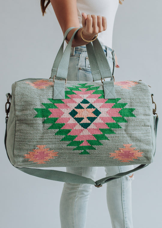 A woman in light blue jeans and a white top holds the panacheaccessories Hadley Duffel, featuring pink and green western-inspired geometric patterns, cotton lining, two handles, a shoulder strap, and a secure zipper closure.