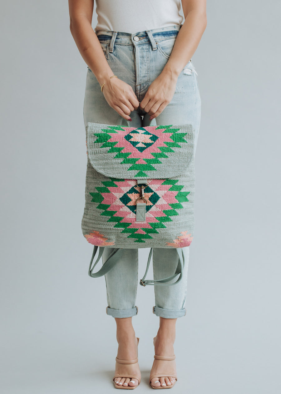 A person in light-wash jeans, white top, and beige heels holds the panacheaccessories Hadley Backpack, featuring green, pink, and black geometric patterns with adjustable straps. The colorful bag is set against a light gray background.