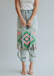 A woman in a white sleeveless top, light blue high-waisted jeans, and beige heels holds the panacheaccessories Hadley Backpack—a colorful, geometric-patterned western-inspired bag with green, pink, and black designs—in front of her.
