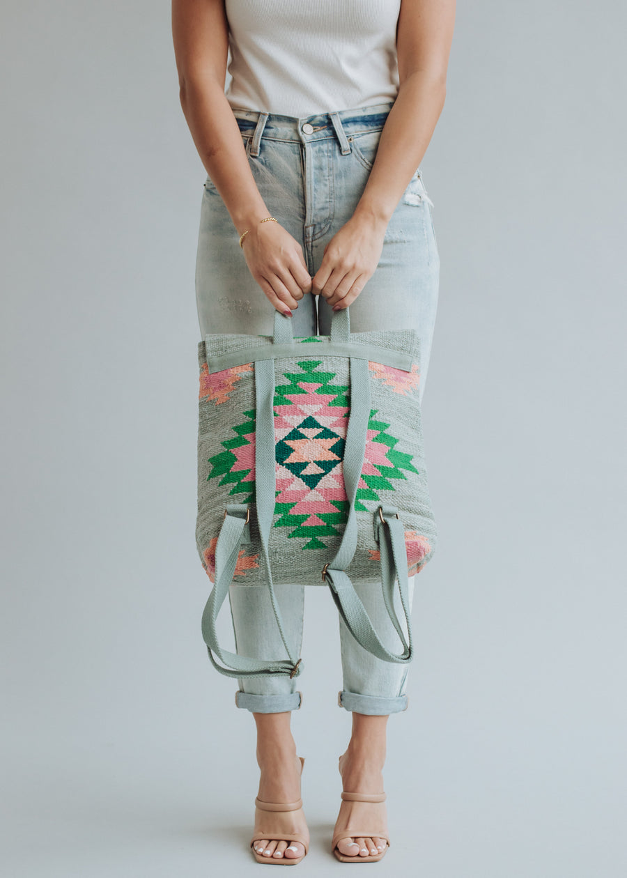 A woman in a white sleeveless top, light blue high-waisted jeans, and beige heels holds the panacheaccessories Hadley Backpack—a colorful, geometric-patterned western-inspired bag with green, pink, and black designs—in front of her.