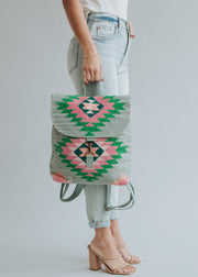 A woman in light-wash jeans and nude heeled sandals holds the panacheaccessories Hadley Backpack, featuring a green, pink, and black geometric pattern with adjustable straps, against a light gray background.