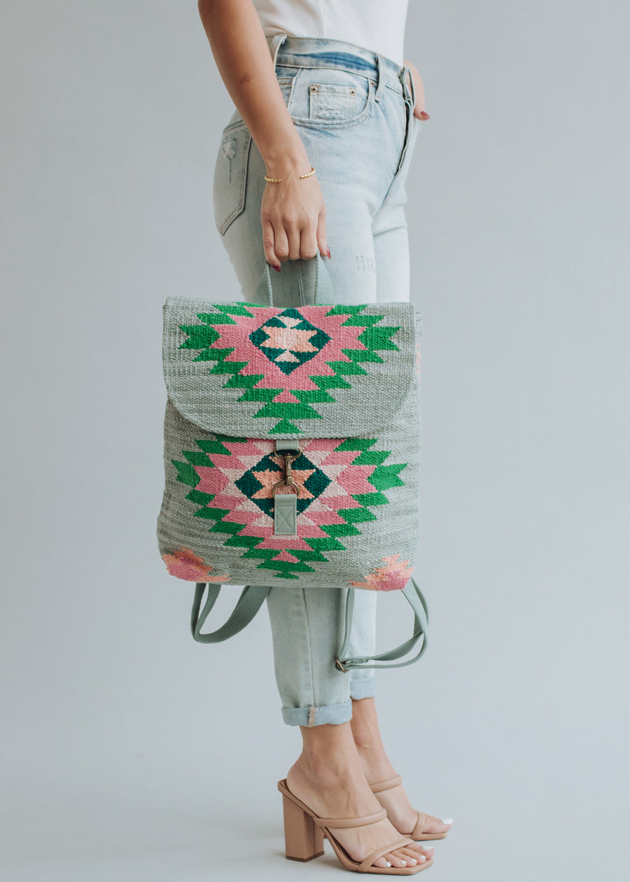 A woman in light-wash jeans and nude heeled sandals holds the panacheaccessories Hadley Backpack, featuring a green, pink, and black geometric pattern with adjustable straps, against a light gray background.