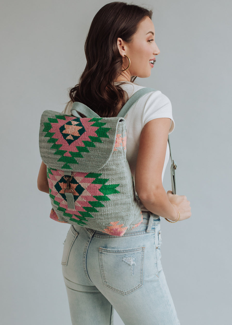 A woman with long brown hair, dressed in a white t-shirt and light blue jeans, smiles sideways while wearing the panacheaccessories Hadley Backpack—western-inspired with green, pink, and gray geometric patterns and adjustable straps.