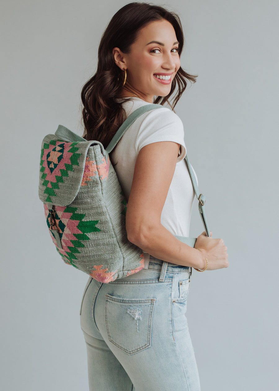 A woman in a white top and light blue jeans smiles over her shoulder, showcasing the panacheaccessories Hadley Backpack with green, pink, and black geometric patterns and adjustable straps, set against a plain light gray background.