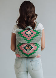 A woman with wavy brown hair, in a white shirt and light blue jeans, stands facing away, showing the panacheaccessories Hadley Backpack featuring adjustable straps and a pink and green geometric western-inspired pattern.