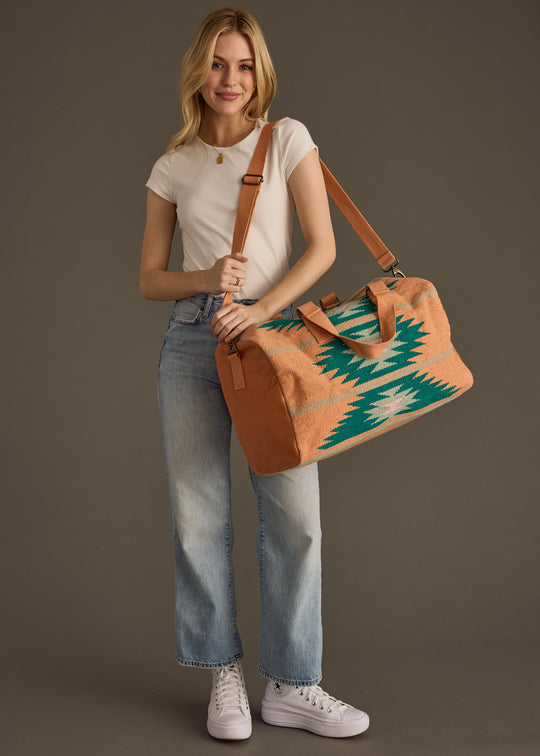 A woman in a white t-shirt, light blue jeans, and white sneakers smiles while holding the panacheaccessories Quinn Duffel, a large western-inspired bag with green and white geometric patterns and a secure zipper, against a brown background.