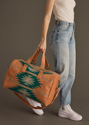 A person in a white sleeveless top, light blue jeans, and white high-top sneakers holds the panacheaccessories Quinn Duffel, featuring a green and blue geometric pattern with zipper closure, against a gray background.