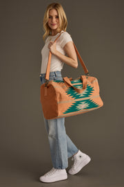 A smiling woman in a white t-shirt, blue jeans, and white sneakers carries the Quinn Duffel by panacheaccessories—featuring leather straps and a zipper closure—over her shoulder against a plain background.