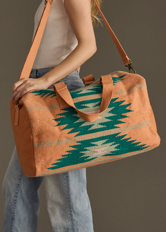 A person wearing a white sleeveless top and blue jeans carries the panacheaccessories Quinn Duffel, featuring orange and teal geometric designs, tan straps, zipper closure, and interior pockets, over their shoulder.