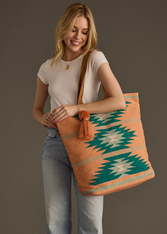 A smiling woman in a white t-shirt and light jeans holds the Quinn Tote by panacheaccessories, a large western-inspired bag with a leather suede shoulder strap and tassel, standing against a plain brown background.