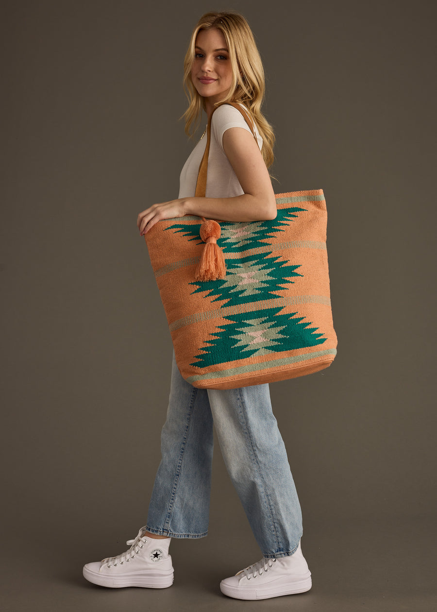 A woman stands sideways, smiling, wearing a white t-shirt, light blue jeans, and white sneakers. She carries the panacheaccessories Quinn Tote—an orange and teal western-inspired bag with a leather suede shoulder strap and tassel.