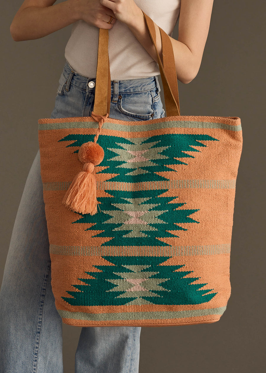 A person in a white sleeveless top and blue jeans holds the panacheaccessories Quinn Tote, a western-inspired bag with a geometric teal, white, and peach pattern, tan handles, snap closure, plus an orange tassel and pom-pom.
