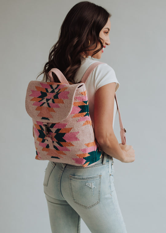 A woman in a white t-shirt and light-wash jeans models the panacheaccessories Mari Backpack, featuring a pink and multicolored geometric pattern and adjustable straps, against a plain light background.