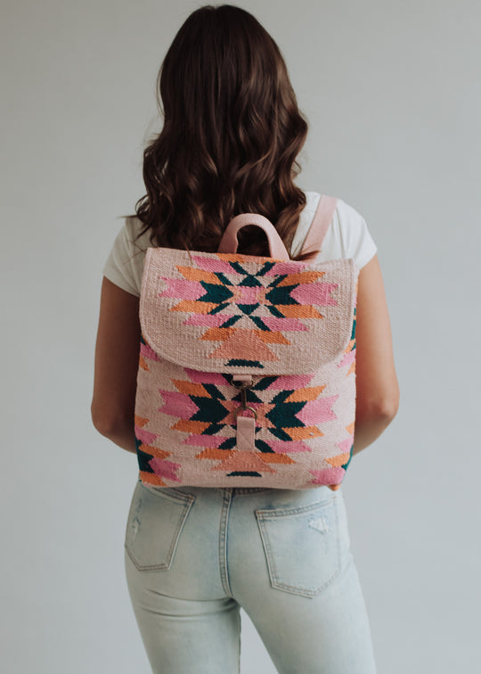 A woman with long brown hair, in a white t-shirt and light jeans, stands facing away, showcasing the Mari Backpack by panacheaccessories—a pink western bag with a colorful geometric pattern and adjustable straps.