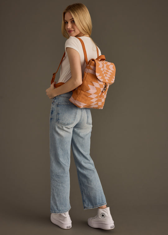 A woman faces backward, turning her head to the camera. She wears a white t-shirt, light blue jeans, white Converse sneakers, and the tan Elisa Backpack from panacheaccessories featuring adjustable straps and a pink geometric pattern.